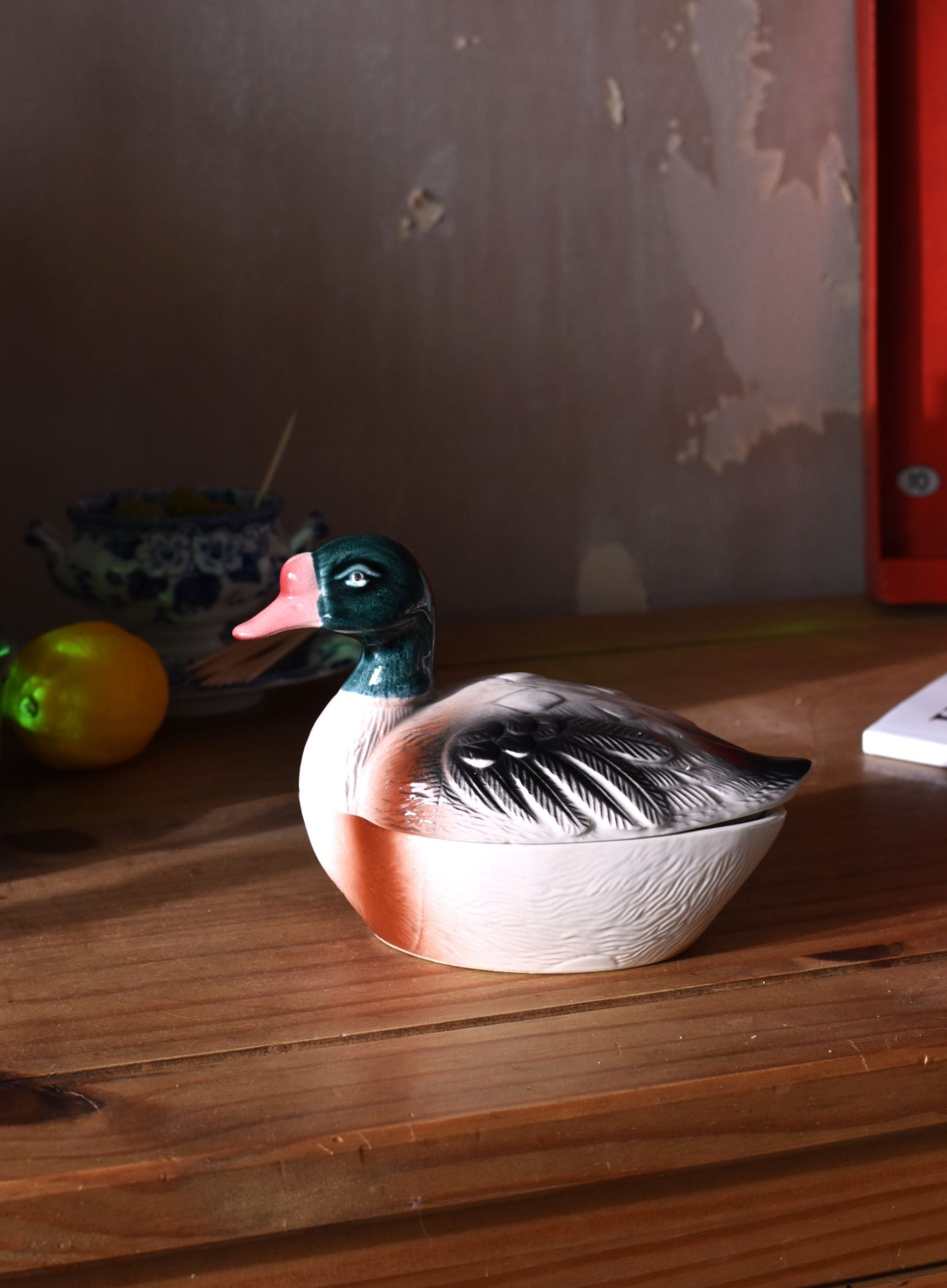 a covered dish in the shape of a duck, painted grey and green, on a wooden surface