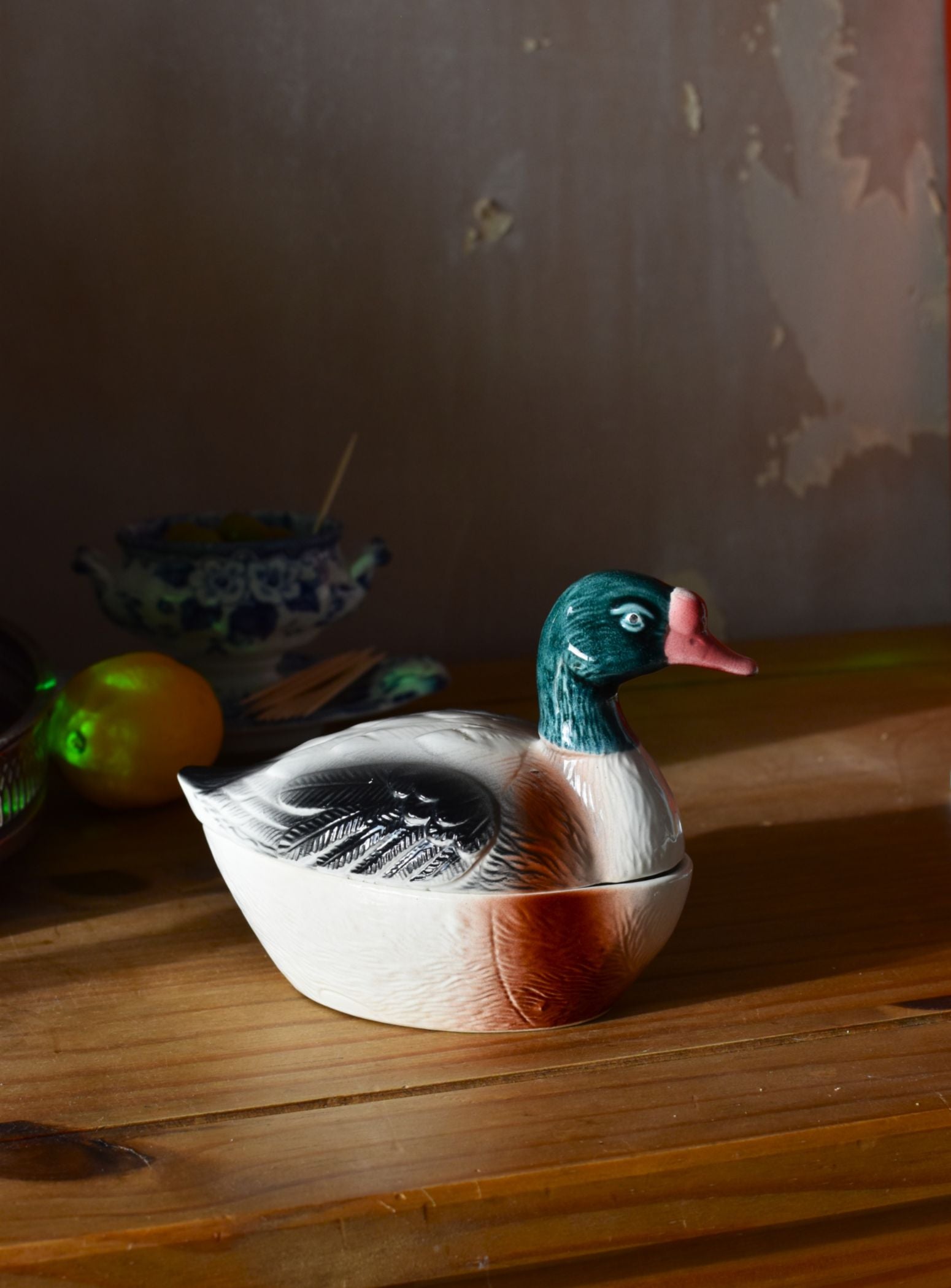a covered dish in the shape of a duck, painted grey and green, on a wooden surface