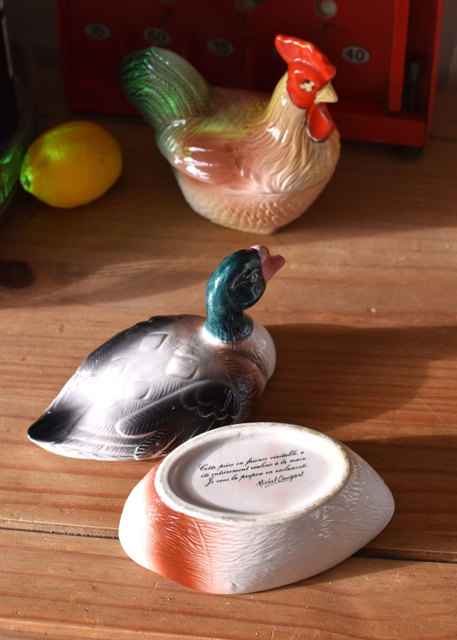 a covered dish in the shape of a duck, painted grey and green, on a wooden surface, with a cockerel in the background