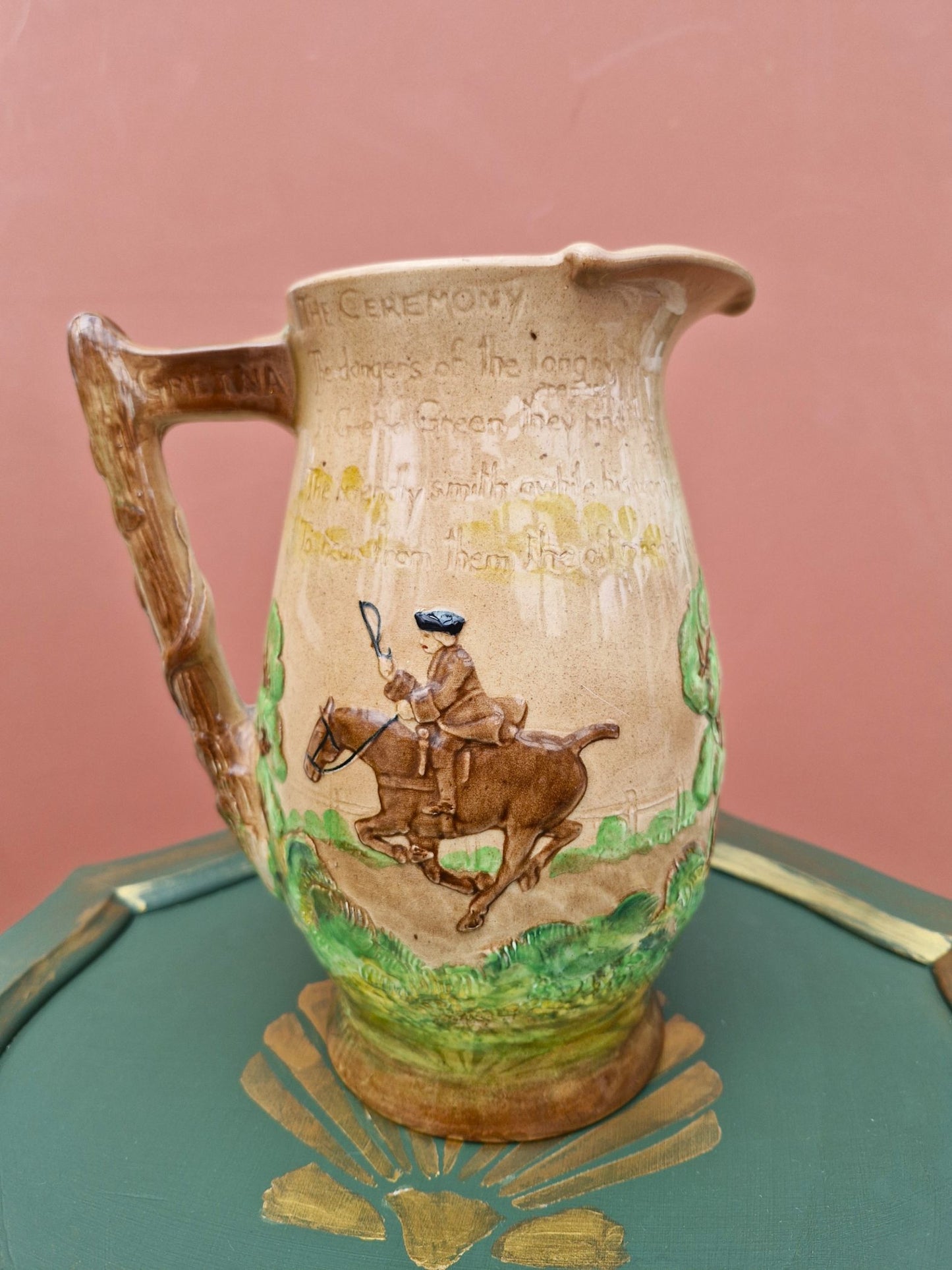 Ceramic pitcher with a decorative design of a person on horseback, placed on a green surface with a pink background.