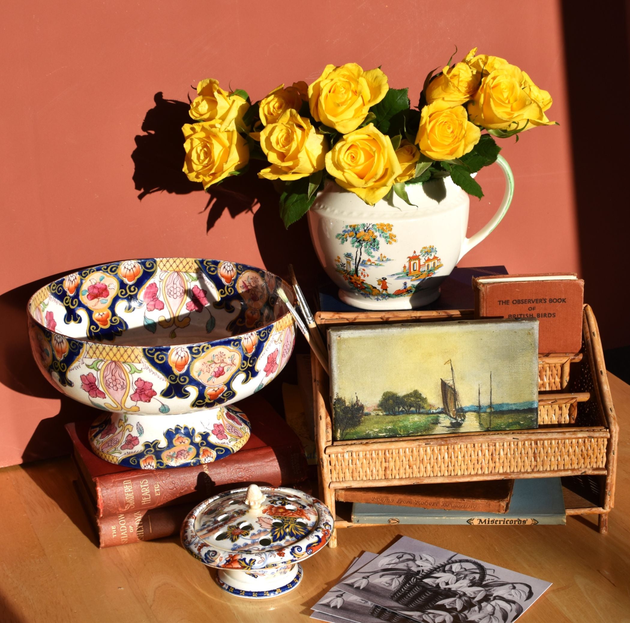Decorative items including a floral teapot, bowl, and books on a wooden surface with a pinkish-brown background.
