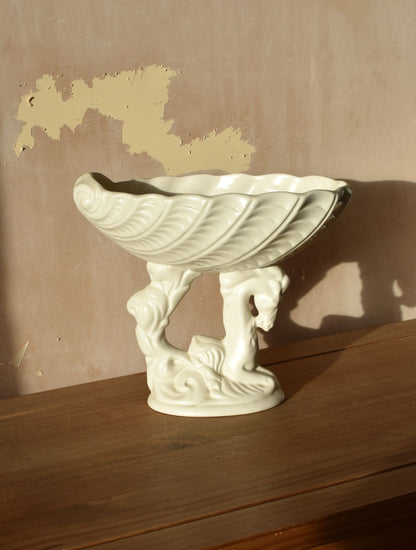 a white ceramic vase in the shape of a shell on top of a seahorse on a wooden surface