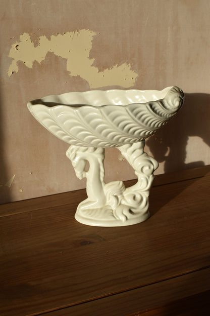a white ceramic vase in the shape of a shell on top of a seahorse on a wooden surface