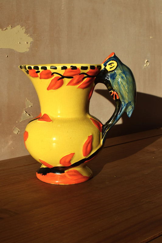 a yellow and orange art deco ceramic jug with a blue parrot shaped handle against a neutral background