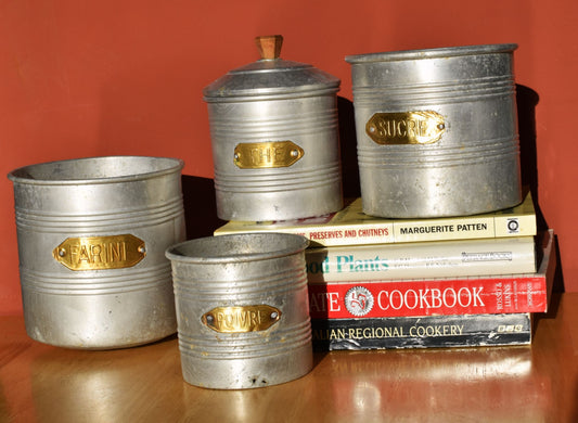 four silver tins with gold labels are stacked on a table and some books against a dark red wall
